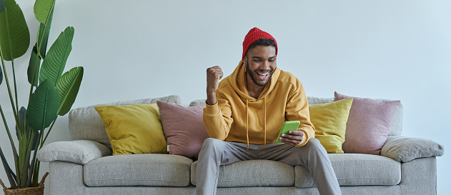 Happy young African man using smart phone and gesturing while sitting on the couch