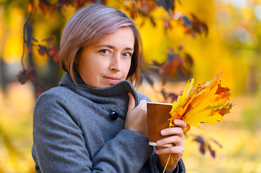 portrait of a beautiful woman with paper cup and colorful yellow leaves, posing in a city park, bright sunny day in autumn
