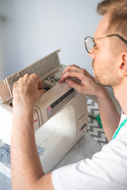 istock A blond man with glasses changes a bobbin in a sewing machine. A tailor with a measuring tape on his neck in a white t-shirt. The concept of small business, lifestyle and enjoying of work 1406515361