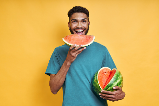 Happy African man eating watermelon while standing against yellow background