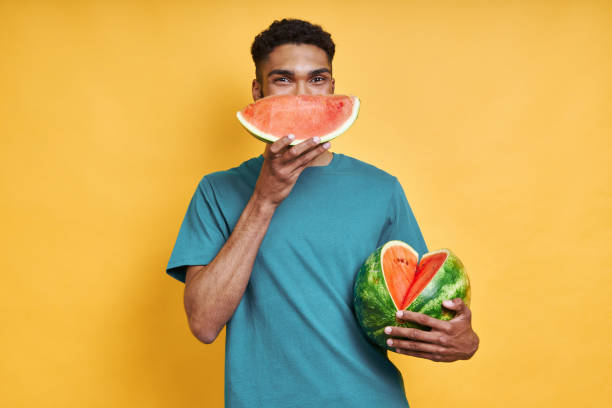 Playful African man holding watermelon Playful African man holding watermelon while standing against yellow background black man eating watermelon stock pictures, royalty-free photos & images
