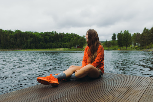 Happy female in orange with long hair enjoying the scenic Swedish nature, relaxing on the wooden pier with view of the lake surrounded by the green pine forest