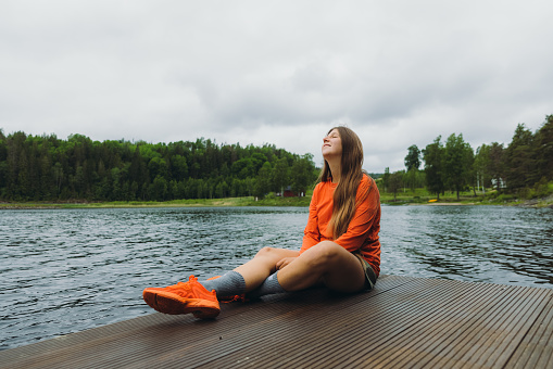 Happy female in orange with long hair enjoying the scenic Swedish nature, relaxing on the wooden pier with view of the lake surrounded by the green pine forest