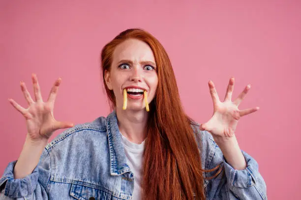 funny redhaired ginger woman with potatoes like canine tooth scaring, her hands want grab you in studio pink background. fright candies or life concept.