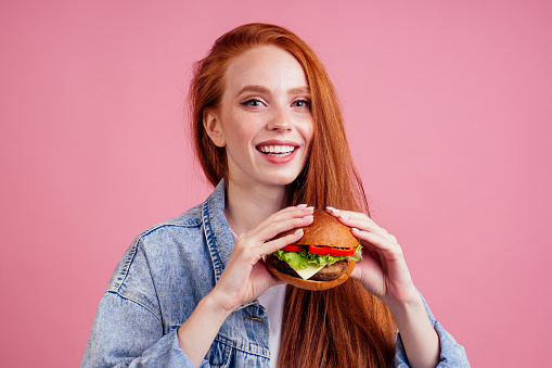 redhaired ginger woman with freckles enjoying big huge burger cutlet and wearing demin american jeans jacket in studio pink background .USA traditional concept.