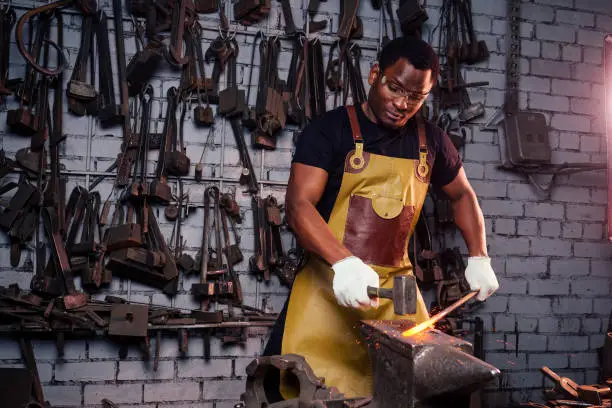 handsome african americam man forging steel next to furnace in dark workshop. small business comcept