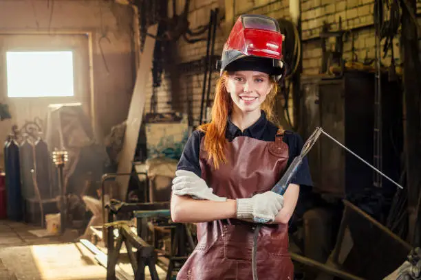 Strong and worthy pretty redhead ginher woman wearing protection helmet and leather apron with gloves holding welding machine instrument . small business concept.