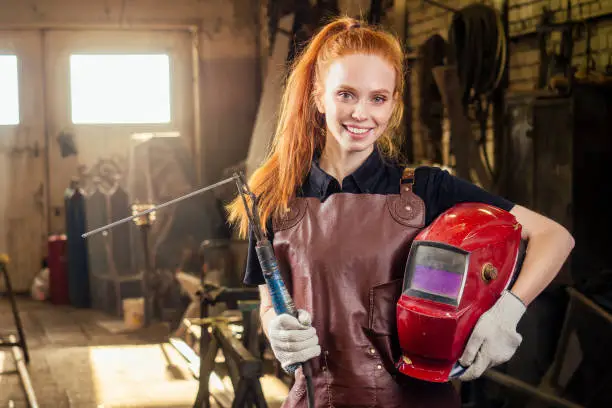 Strong and worthy pretty redhead ginher woman wearing protection helmet and leather apron with gloves holding welding machine instrument . small business concept.