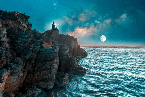 Photo of silhouette of a person meditating on cliff at night with milky way over the sea