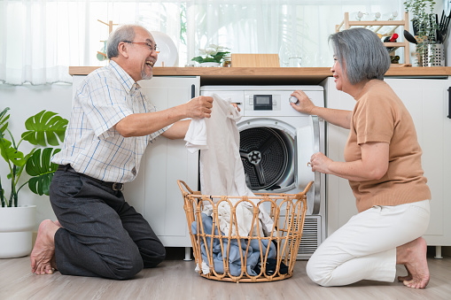 Happy Asian senior older man helping his wife put clothes in washing machine in laundry room