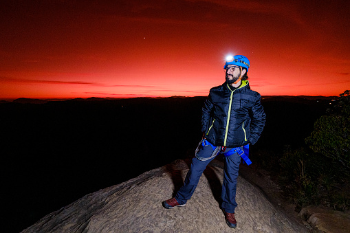 Young latin man with beard on mountain peak with safety equipment at sunrise.
