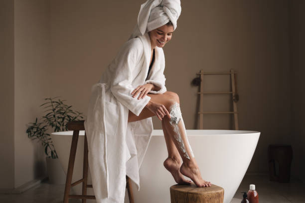 istock Woman depilating legs using shaving foam and razor, dressed in bathrobe standing in bathroom 1406542046