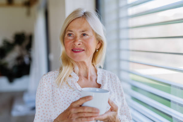 istock Attractive senior woman drinking tea and standing by window at home. 1406548452