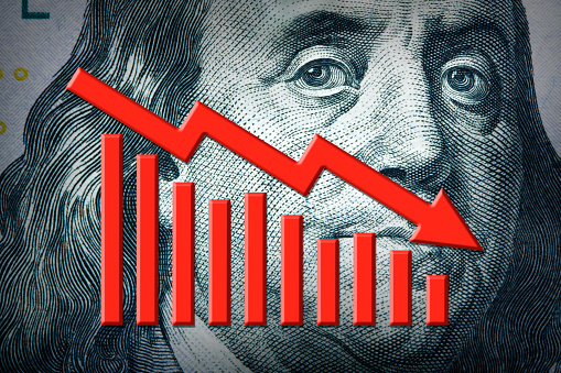 A close up of a red declining bar chart and arrow positioned on the face of Benjamin Franklin on a U.S. one hundred dollar bill.