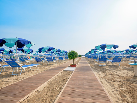 Boardwalk on the beach along a row of colorful umbrellas and sunbeds leading to the sea on a sunny summer day on the shore of the Adriatic Sea, Italy. Early morning sun on the beach.