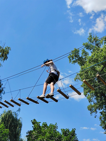 A teenager boy walks along a rope bridge between trees in an amusement park in safety gear and a helmet. Photo from the bottom