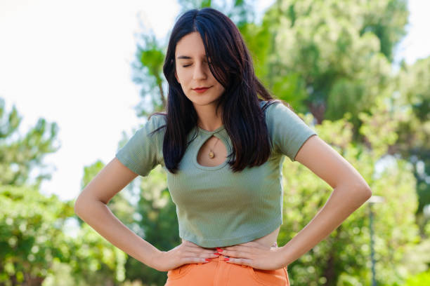 Brunette millennial woman wearing turquoise tee on city park, outdoors with hand on stomach because indigestion, painful illness feeling unwell. Ache concept. stock photo