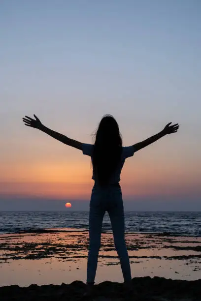 Photo of Teen woman looking to sunset, rejoicing by raising her hands up against the sea in israel. Triumph silhouette