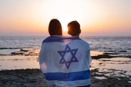 Teenagers, young women and man with the Flag of Israel draped over their shoulders at the sunset over the sea in israel. Friendship in childhood silhouette