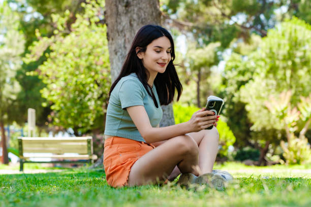 Young brunette girl smiling happy wearing turquoise t-shirt standing on city park, outdoors holding takeaway coffee mug and using mobile smart phone. Scrolling on social media or messaging. stock photo