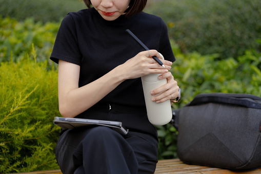 Young Asian freelancer drinking water from reusable bottle during working while sitting in a bench near electric scooter at office park.
