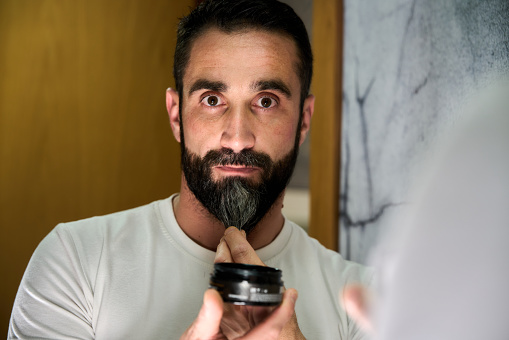 Young man applying wax and oil to his beard in front of mirror. High quality photo