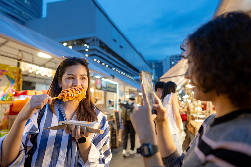 Asian influencer eat food while taking a selfie video with friend at night food market.