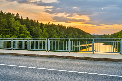 Photo of a road going over a bridge over a river at sunset with a beautiful orange cloudscape and forest in the background.