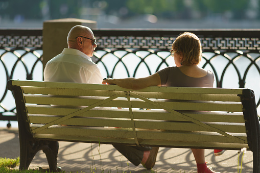 Moscow, Russia - June 10, 2022: Family of mature man and woman sitting on the bench in front of Moskva river in the public park. Time to rest and talking. Concept of privacy. Back. rear view