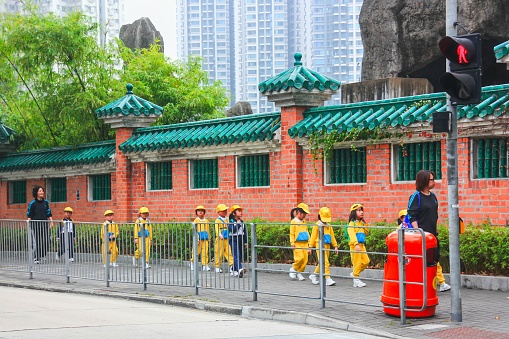 Chinese young students of elementary school wearing yellow school uniform are going to excursion with teachers along ancient brick Buddhist temple wall. Hong Kong,China,Feb 2013