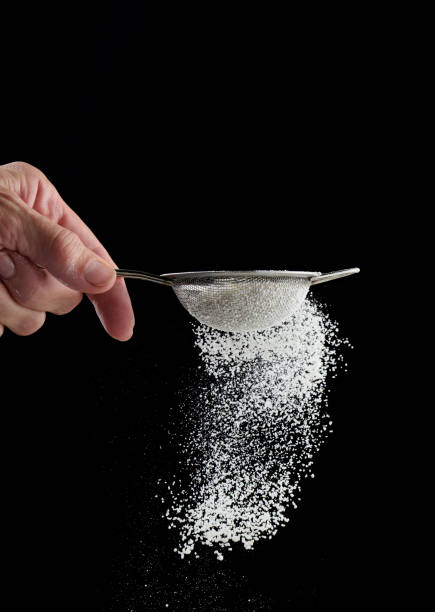 Female hand holds a metal strainer with powdered sugar. Particles fly in different directions Female hand holds a metal strainer with powdered sugar. Particles fly in different directions on a black background, sprinkling sifting stock pictures, royalty-free photos & images