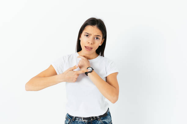 Dissatisfied young woman points at the clock with claim that you were late again, could not complete the task on time, deadline, white studio background