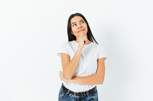 Happy dreaming young woman wearing white T-shirt and blue jeans with her hand under her chin, looking up wishing for something, white studio background