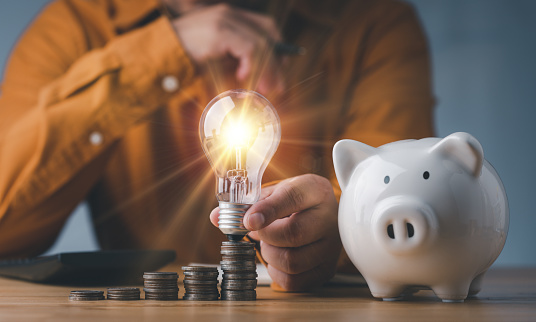saving energy and money concept. idea for save or investment. businessman holding lightbulb on piggy bank and coins stacking on desk with calculator.