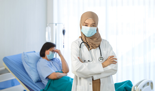 Young muslim female doctor arms crossed stand in hospital with elderly patient lie down on bed on the background