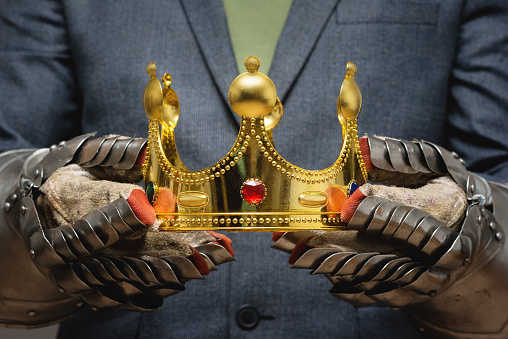 Business success concept. Businessman in the plate armor mittens holds a golden crown close up. Authority power.