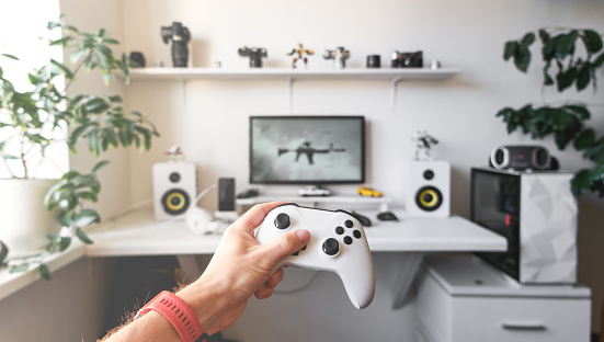 Close-up of male hands holding white wireless gamepad against white computer dream desk with big monitor.