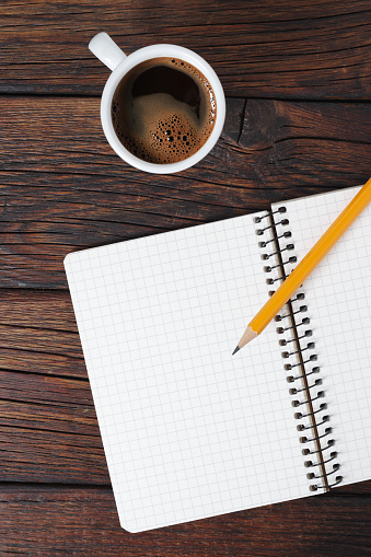 Notepad with pencil and cup of coffee on wooden table, top view