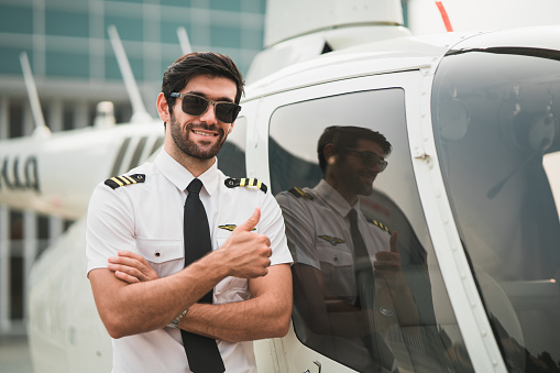 Portrait of optimistic helicopter pilot standing with aircraft and smiling while looking at camera. Good looking and confident man at hangar outdoor. aircraft carrier occupation. Ready to fly.