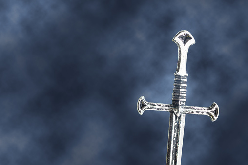 A medieval knight in full suit of armour and embroidered cape kneeling inside a castle in front of two flaming torches leaning on a broad sword in contemplation. With subtle backlight of an overhead shaft of light. With shallow depth of field, focus on the knight.