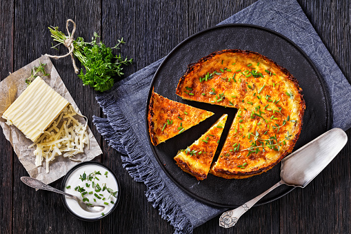 bacon and cheddar quiche with shredded potato crust on black plate with cheddar cheese, sour cream and parsley thyme bouquet on dark wooden table, horizontal view, flat lay
