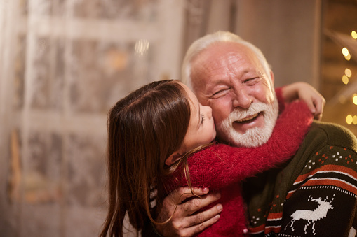 Happy mature man enjoying in a kiss and hug by his small granddaughter at home on Christmas day.