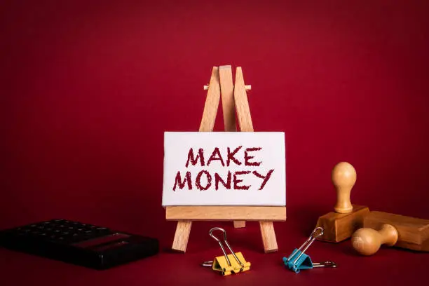 Photo of Make Money. Miniature easel with text and office supplies on red background