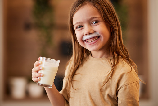 Happy little baby girl smiles happily when drinking delicious milk from a glass sitting at a table in the kitchen at home.
