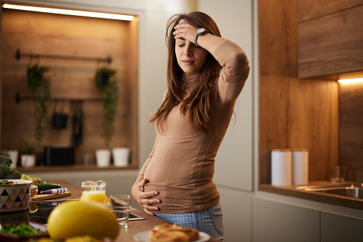 Young pregnant woman holding her head in pain in the kitchen.