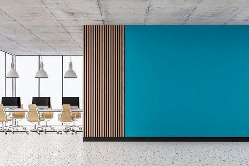 Empty office interior with a large empty azure blue colored plaster wall and partly hardwood paneled wall with copy space on the terrazzo floor. Work desks, beige chairs, lighting equipment, computer equipment, and windows in the background. 3D rendered image.