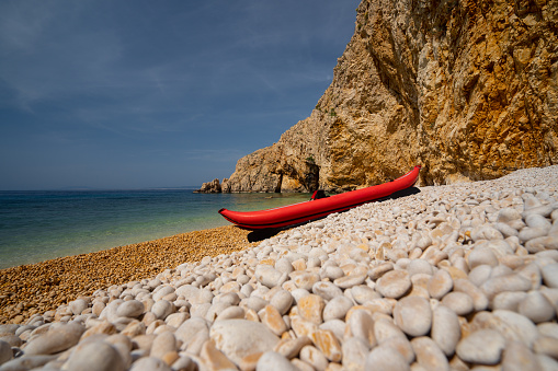 Red kayak on deserted shingle beach of beautiful croatian island with cliffs and calm sea on sunny day