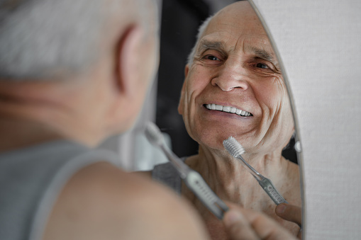 Smiling old man brushing his teeth prosthesis with toothbrush. Odontic healthcare concept.