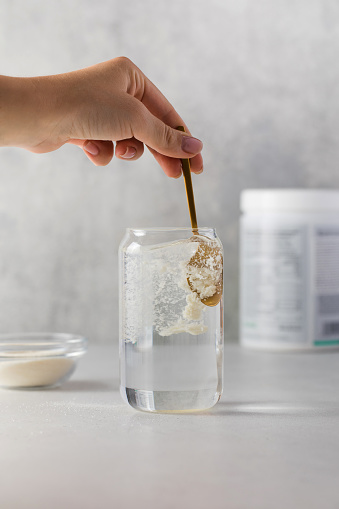 Hydrolyzed collagen powder is added with a spoon to a transparent glass of water on a grey background. The concept of health, anti-aging dietary supplements.
