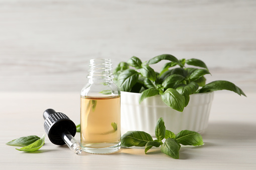 Glass bottle of basil essential oil near bowl with leaves on white wooden table